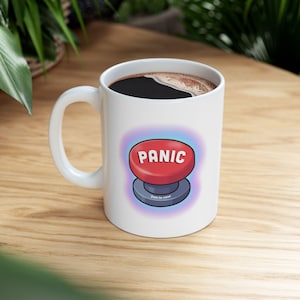 Peut inclure: Mug en céramique blanche avec une anse, rempli de liquide foncé. Le mug présente un bouton rouge avec le mot "PANIC" en lettres blanches. En dessous, le texte indique "Just in case".