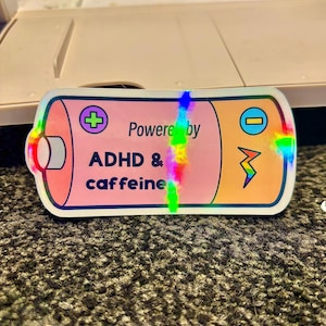 Könnte beinhalten: Ein holografischer Aufkleber in Form einer Batterie mit rosa und orangefarbenen Abschnitten. Der Aufkleber lautet "Powered by ADHD & caffeine" mit einem Pluszeichen, einem Minuszeichen und einem Blitz.