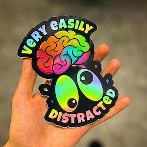 May include: A holographic sticker with the words "VERY EASILY DISTRACTED" in a rainbow gradient. The sticker features a colourful brain and large, cartoonish eyes.
