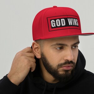Puede incluir: Una gorra de béisbol roja y negra con el texto "GOD WINS" en letras blancas. La gorra tiene una visera plana y una correa negra.