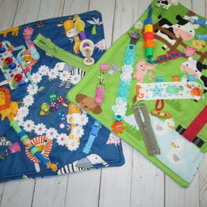 May include: Two colorful sensory activity mats for babies. One is blue with animal graphics and various attachments. The other is green with farm animal graphics and different textures and fasteners. Each mat is designed to stimulate sensory exploration.