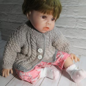 May include: A doll wearing a gray cable-knit cardigan with two white buttons. The doll is also wearing pink floral pants and white socks with pink accents. The doll has brown eyes and brown hair.