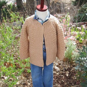May include: A tan, hand-knitted cardigan sweater with a textured stitch pattern. The open-front sweater is displayed on a mannequin, layered over a plaid shirt and blue jeans. The sweater has long sleeves and a classic, casual design.