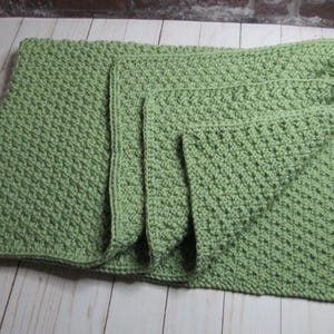 May include: A light green, handmade crochet blanket with a textured stitch pattern. The blanket is folded, revealing a layered design. The soft, muted green color and the cozy texture suggest a handmade item, perfect for home decor or as a baby blanket.
