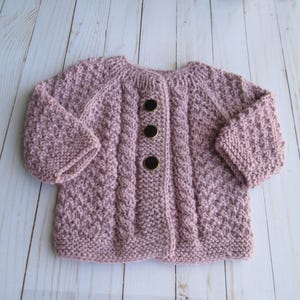 May include: A light mauve knitted cardigan with three dark brown buttons. The baby sweater features a textured stitch pattern and cable knit detailing down the front. The sleeves are three-quarter length, and the garment is made of soft yarn.
