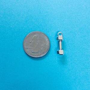 May include: A silver dumbbell charm next to a US quarter for size comparison. The charm is a simple design with a rounded top and bottom.