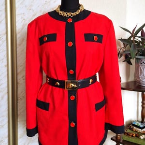Könnte beinhalten: Rote Jacke mit schwarzen Verzierungen, Taschen und Knöpfen. Die Jacke wird mit einem schwarzen Gürtel mit goldener Schnalle in der Taille zusammengehalten. Eine goldene Halskette liegt um den Hals der Büste.