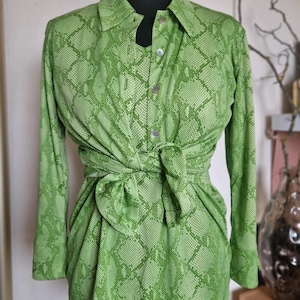 Puede incluir: Una camisa de manga larga con botones en un vibrante color verde lima con un patrón de piel de serpiente repetitivo. La camisa está atada a la cintura con un nudo, creando un efecto fruncido. Tiene un cuello clásico y botones visibles.