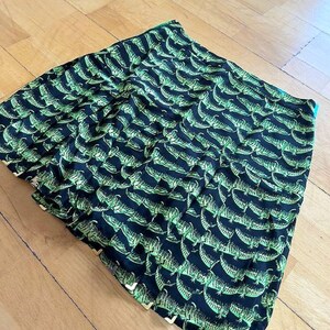 May include: A folded black fabric with a repeating pattern of green alligators. The fabric has a slight sheen and appears to be a scarf or a similar accessory. The alligator design is detailed and covers the entire surface.