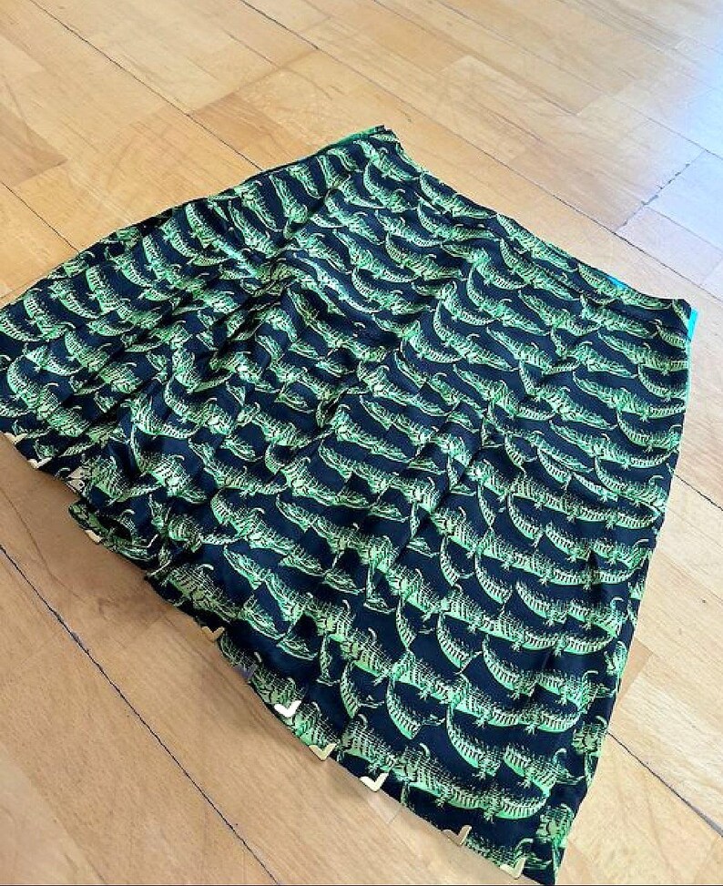 May include: A folded black fabric with a repeating pattern of green alligators. The fabric has a slight sheen and appears to be a scarf or a similar accessory. The alligator design is detailed and covers the entire surface.