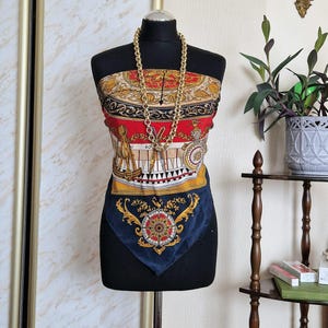 May include: A silk scarf in red, gold, and navy blue, displayed on a black mannequin. The scarf has intricate patterns and a gold chain necklace. The text "natural silk" is visible at the bottom. The scarf is likely around 70cm x 70cm.