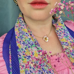 May include: A gold necklace with a flower pendant is worn over a pink top. A colourful floral scarf and gold earrings are also visible. Pink and white flowers are held near the face.