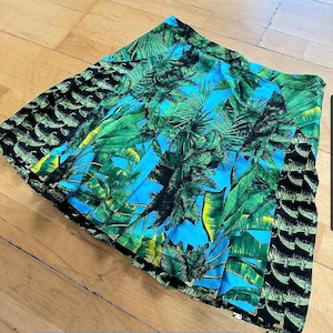May include: A pleated skirt with a vibrant tropical print. The skirt features a design of green palm leaves and blue sky, with a black crocodile pattern on the sides. The text "Versace 100% silk" is visible.