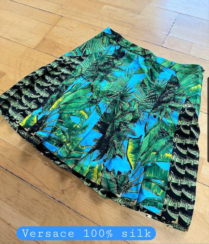 May include: A pleated skirt with a vibrant tropical print. The skirt features a design of green palm leaves and blue sky, with a black crocodile pattern on the sides. The text "Versace 100% silk" is visible.