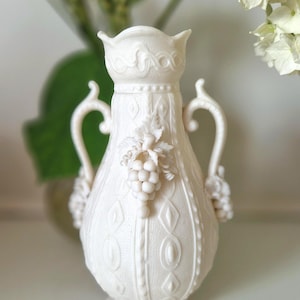 Può includere: Un vaso in ceramica bianco opaco con due manici curvi e una superficie testurizzata con motivi geometrici in rilievo. Il design centrale include un rilievo tridimensionale di grappoli d'uva e foglie sulla parte anteriore e sui lati.
