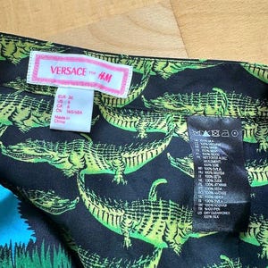 May include: Close-up of a black silk fabric with a repeating pattern of green alligators. The fabric has a tag that reads "VERSACE for H&M" and includes size information. The fabric is 100% silk.