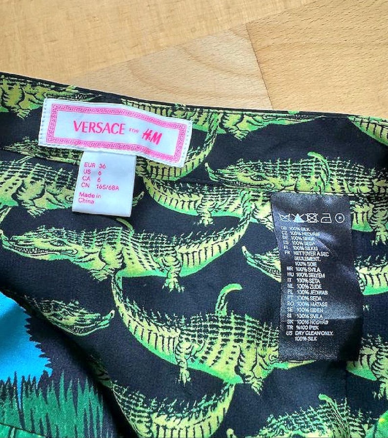 May include: Close-up of a black silk fabric with a repeating pattern of green alligators. The fabric has a tag that reads "VERSACE for H&M" and includes size information. The fabric is 100% silk.