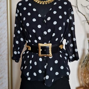 Op de afbeelding: Een zwarte blouse met witte stippen, driekwartmouwen en een V-hals. De blouse is in de taille getailleerd met een brede zwarte leren riem met een grote, getextureerde goudkleurige vierkante gesp en gouden olifantaccenten als versiering.