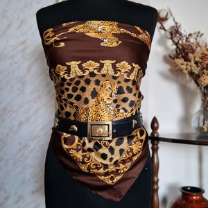 May include: A brown and gold scarf featuring a leopard print and gold detailing, displayed on a black mannequin. A black belt with a gold buckle is worn around the waist. The scarf is square in shape.