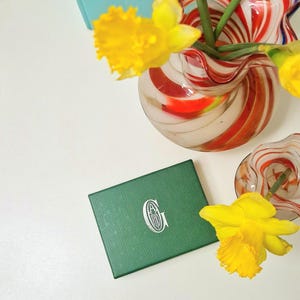 Puede incluir: Una caja verde con un monograma blanco, junto a un jarrón con remolinos rojos y blancos y narcisos amarillos. El jarrón contiene tallos verdes. También hay otro jarrón más pequeño con remolinos rojos y blancos.