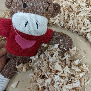 May include: A handmade sock monkey doll with a red sweater and a pink heart applique. The monkey has brown and white knitted details, a white face, and black stitched eyes and mouth. The doll is surrounded by wood shavings.
