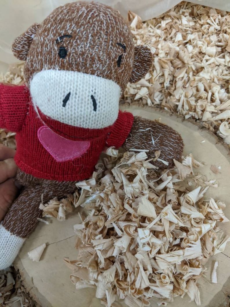 May include: A handmade sock monkey doll with a red sweater and a pink heart applique. The monkey has brown and white knitted details, a white face, and black stitched eyes and mouth. The doll is surrounded by wood shavings.