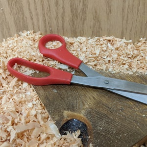 May include: A pair of scissors with red handles and silver blades rests on a wooden surface covered in wood shavings. The scissors are open, ready for use. The background is a light brown wooden surface.