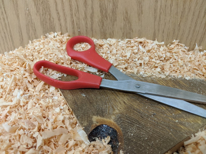 May include: A pair of scissors with red handles and silver blades rests on a wooden surface covered in wood shavings. The scissors are open, ready for use. The background is a light brown wooden surface.