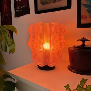May include: A coral-coloured, wavy-edged table lamp emitting a warm glow. The lamp has a black base and sits on a white surface. The lamp's design features vertical lines, creating a textured appearance. The background includes framed artwork and greenery.