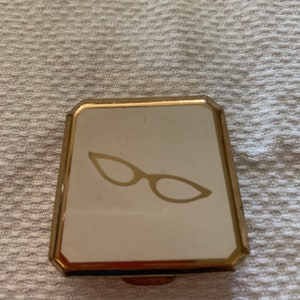 May include: A vintage compact mirror with a gold-tone frame and a white surface. The surface features a gold-colored illustration of eyeglasses. The compact is rectangular with rounded corners.