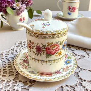 May include: A cream-colored ceramic sugar bowl with a lid and spoon, decorated with floral patterns in red, pink, blue, and yellow. The bowl sits on a matching saucer, with gold trim. A small teacup and vase with flowers are in the background.