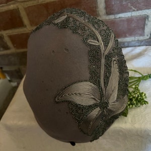 May include: A gray felt hat with a floral design embroidered in silver thread. The hat has a wide brim and a small crown.