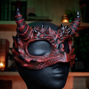 May include: A red and black dragon mask with horns and spiky details. The mask is designed to cover the eyes and upper face, with a textured, scaly appearance. The mask is displayed on a black mannequin head.