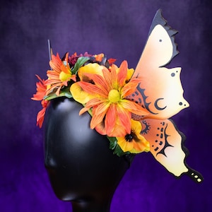 May include: A black mannequin head is adorned with a floral headpiece and a large butterfly wing. The headpiece features orange and yellow flowers. The butterfly wing is orange and cream with black decorative accents.