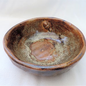 May include: A small, brown ceramic bowl with a speckled glaze. The bowl has a raised acorn design on the rim.