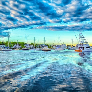 May include: A scenic coastal view featuring a fleet of sailboats and a large white motorboat navigating calm water. The sky is filled with dramatic clouds, and the water reflects the sky's colors. Houses line the shore in the background.