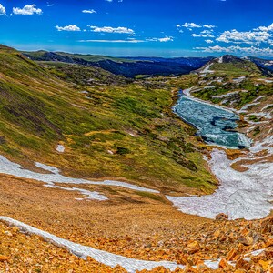 May include: A panoramic view of a mountain range with a winding road leading to a lake. The mountains are covered in snow and the lake is surrounded by green vegetation.