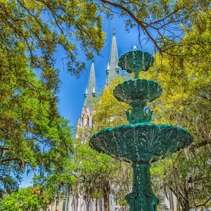 May include: A green fountain with three tiers in a park with tall church spires in the background. The fountain is surrounded by trees with green leaves and some Spanish moss hanging from the branches.