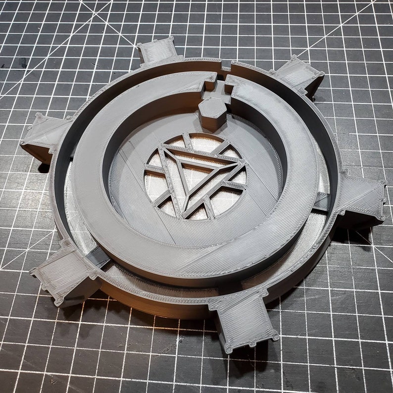 May include: A gray 3D printed model of the Iron Man Arc Reactor. The model is a circular design with a raised center and a detailed Iron Man Arc Reactor design.