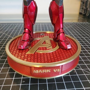 May include: A red and gold action figure stand with a hexagonal pattern and the text "MARK VII". The stand features a gold Avengers logo.