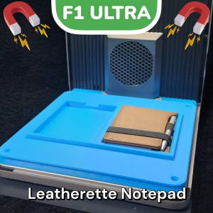 May include: A blue leatherette notepad with a brown leatherette cover and a pen. The notepad is sitting on a gray surface with a black speaker in the background. The text "F1 ULTRA" is visible in green letters above the speaker. The text "Leatherette Notepad" is visible below the notepad.