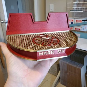 May include: A red and gold 3D printed stand with a circular base and a raised center featuring the Iron Man logo. The stand has the words "MARK VII" inscribed on the front.