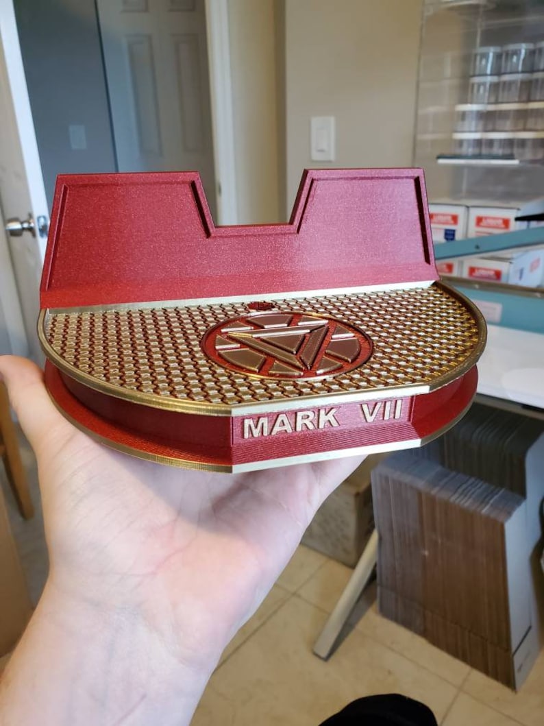 May include: A red and gold 3D printed stand with a circular base and a raised center featuring the Iron Man logo. The stand has the words "MARK VII" inscribed on the front.