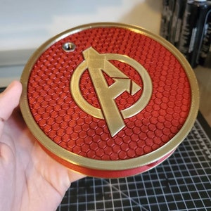 May include: A red and gold 3D printed round wall decor with a honeycomb pattern and the Avengers logo in the center.