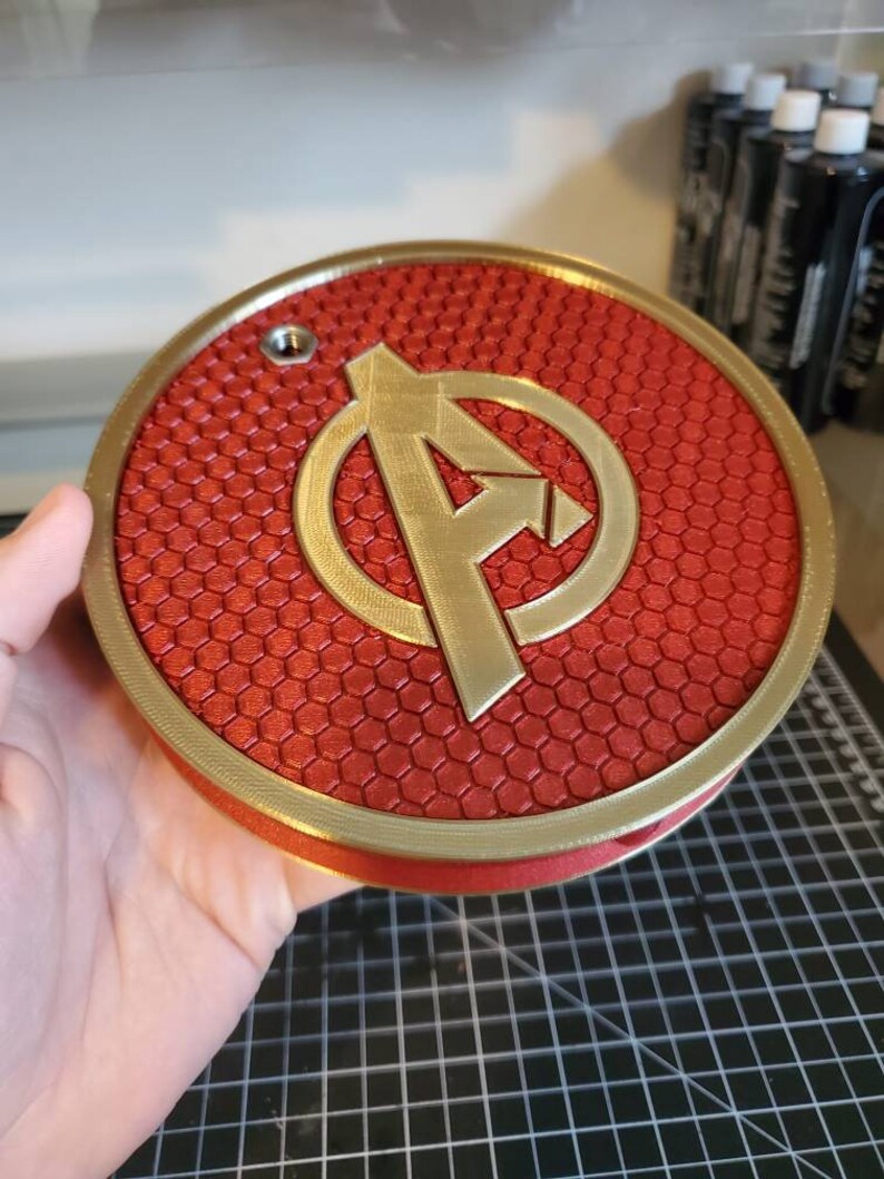 May include: A red and gold 3D printed round wall decor with a honeycomb pattern and the Avengers logo in the center.