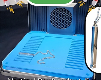 May include: A blue and silver vertical bar pendant jig with a silver bar pendant and chain on the blue surface. The jig has a blue base with a silver border and a blue and silver fan on the back. The text "F1 ULTRA" is written in green letters on the top of the jig. The text "Vertical Bar Pendant Jig" is written in black letters at the bottom of the jig.