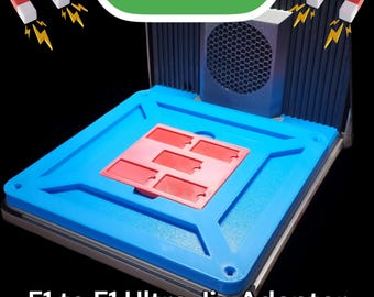 May include: A blue and red 3D printed jig adapter for a 3D printer. The adapter has a rectangular shape with a raised center section. The text "F1 to F1 Ultra Jig Adapter" is printed below the adapter.