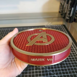 May include: A red and gold circular stand with a honeycomb pattern and the Avengers logo. The stand has the words "MARK VII" inscribed on the bottom.