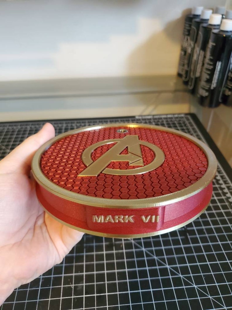 May include: A red and gold circular stand with a honeycomb pattern and the Avengers logo. The stand has the words "MARK VII" inscribed on the bottom.