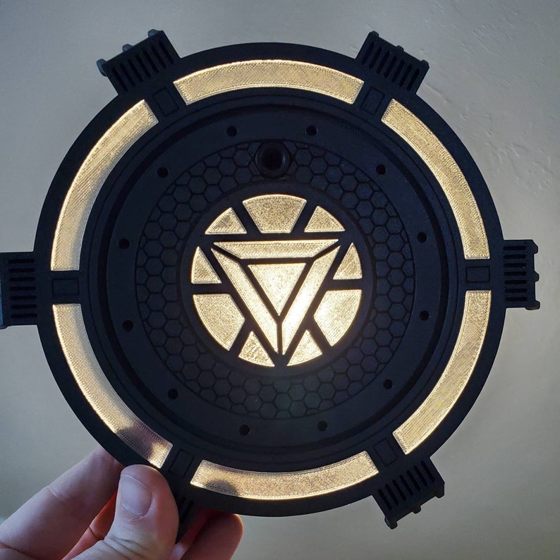 May include: A black and silver circular light fixture with a glowing Iron Man arc reactor design in the center. The light fixture has a textured, honeycomb-like pattern around the reactor.